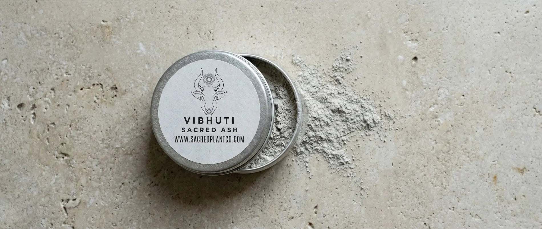 Top-down view of an open tin of Vibhuti Sacred Ash on a textured stone surface, showing the fine grey powder.
