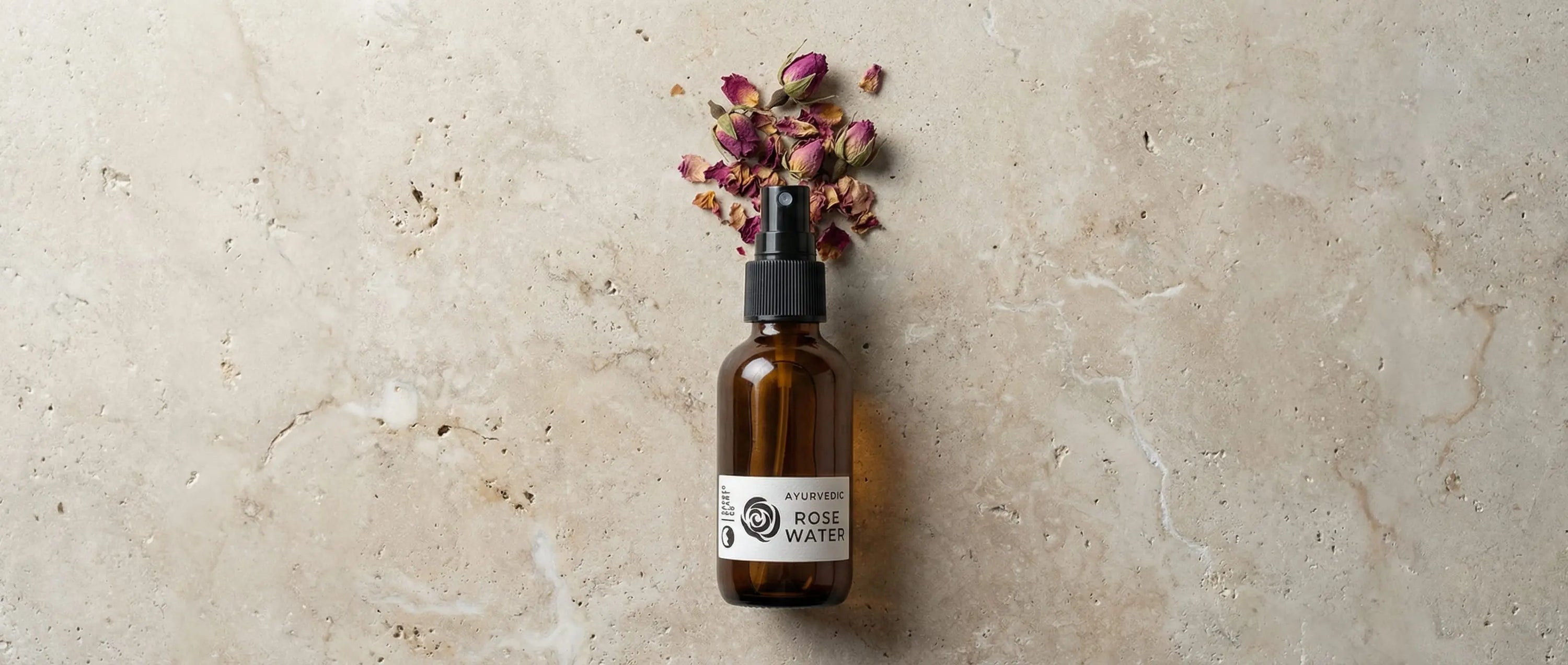 A dark amber glass bottle of Sacred Plant Co Ayurvedic Rose Water lying on a natural stone surface next to spilled dried pink rose buds.