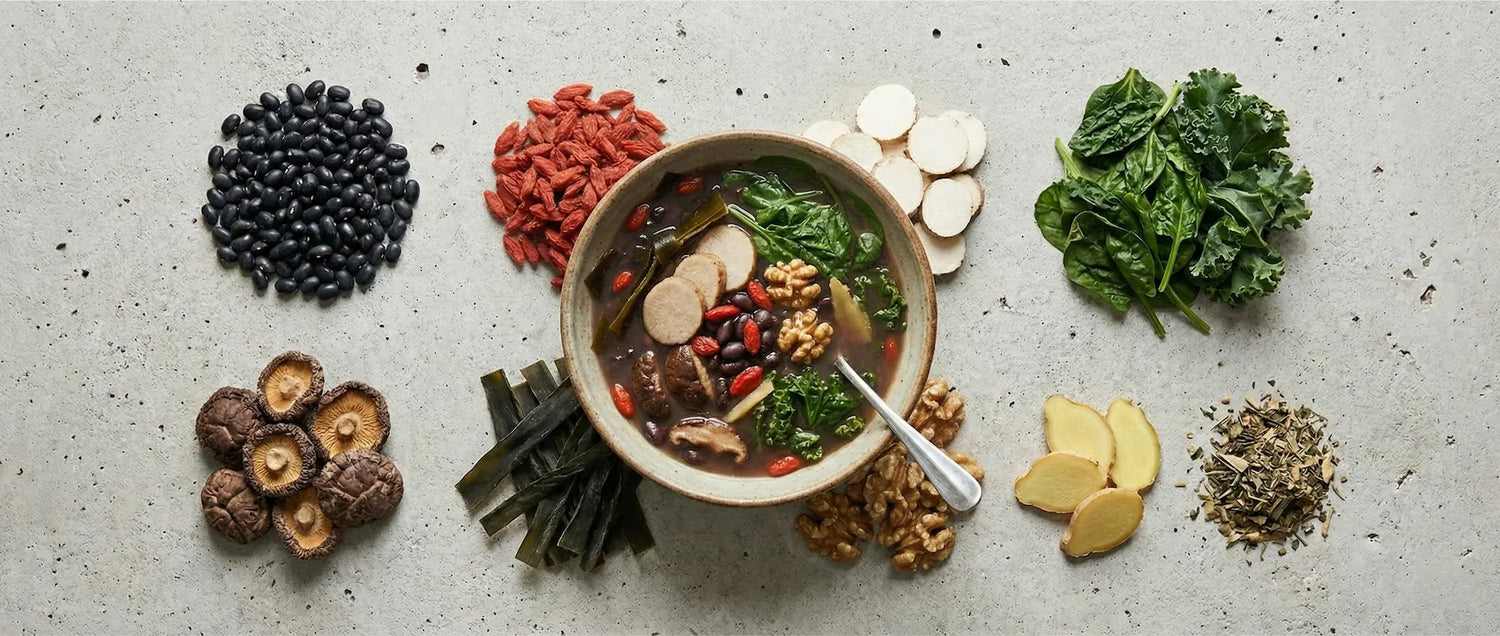 Flat lay arrangement of soup ingredients organized by type: black beans, goji berries, Chinese yam slices, leafy greens, mushrooms, seaweed, ginger, and walnuts surrounding a central bowl.