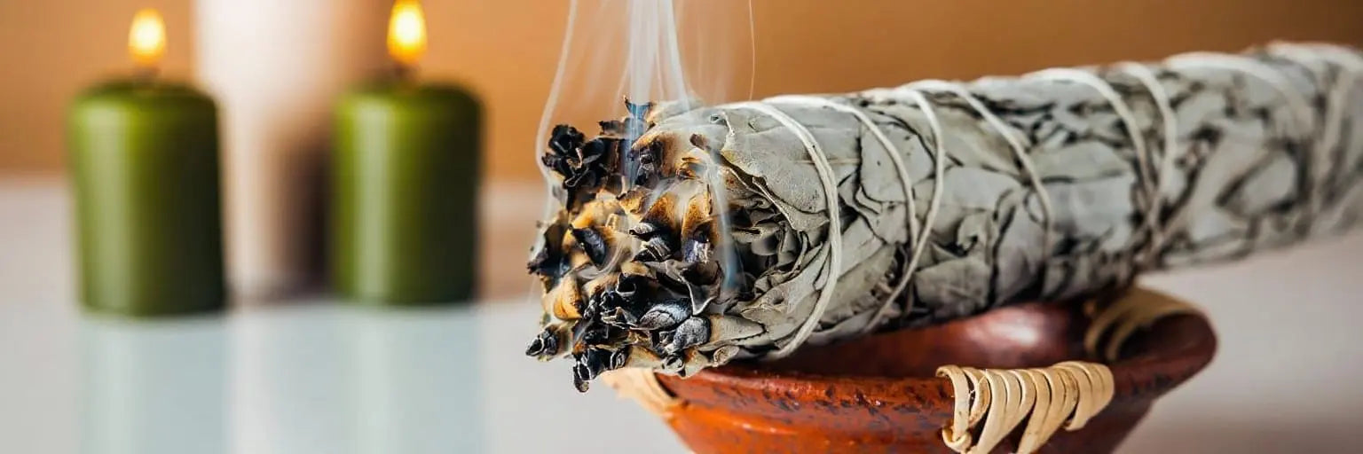 White sage smudge stick burning in a terracotta bowl with smoke rising, surrounded by green candles on a warm wooden surface, depicting a traditional spiritual cleansing and energy purification ceremony.