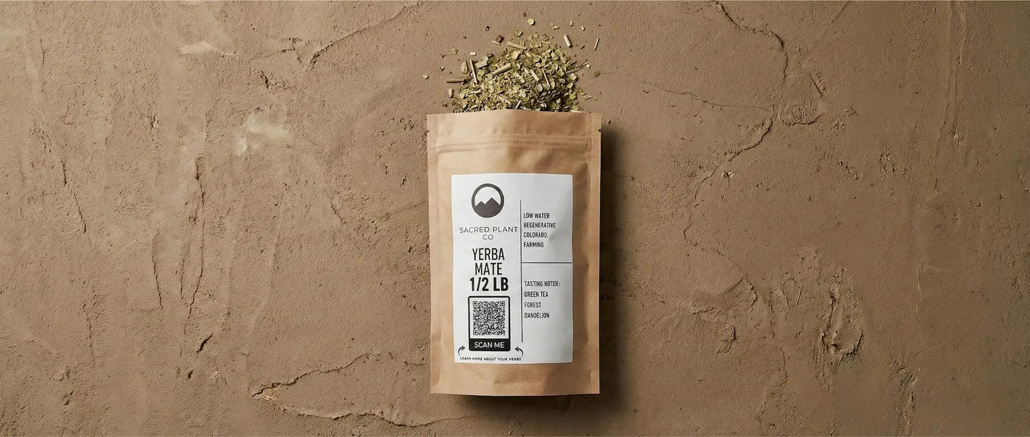 Sacred Plant Co Yerba Mate packaging standing upright with loose dried green leaves spilled on a textured earthy background.