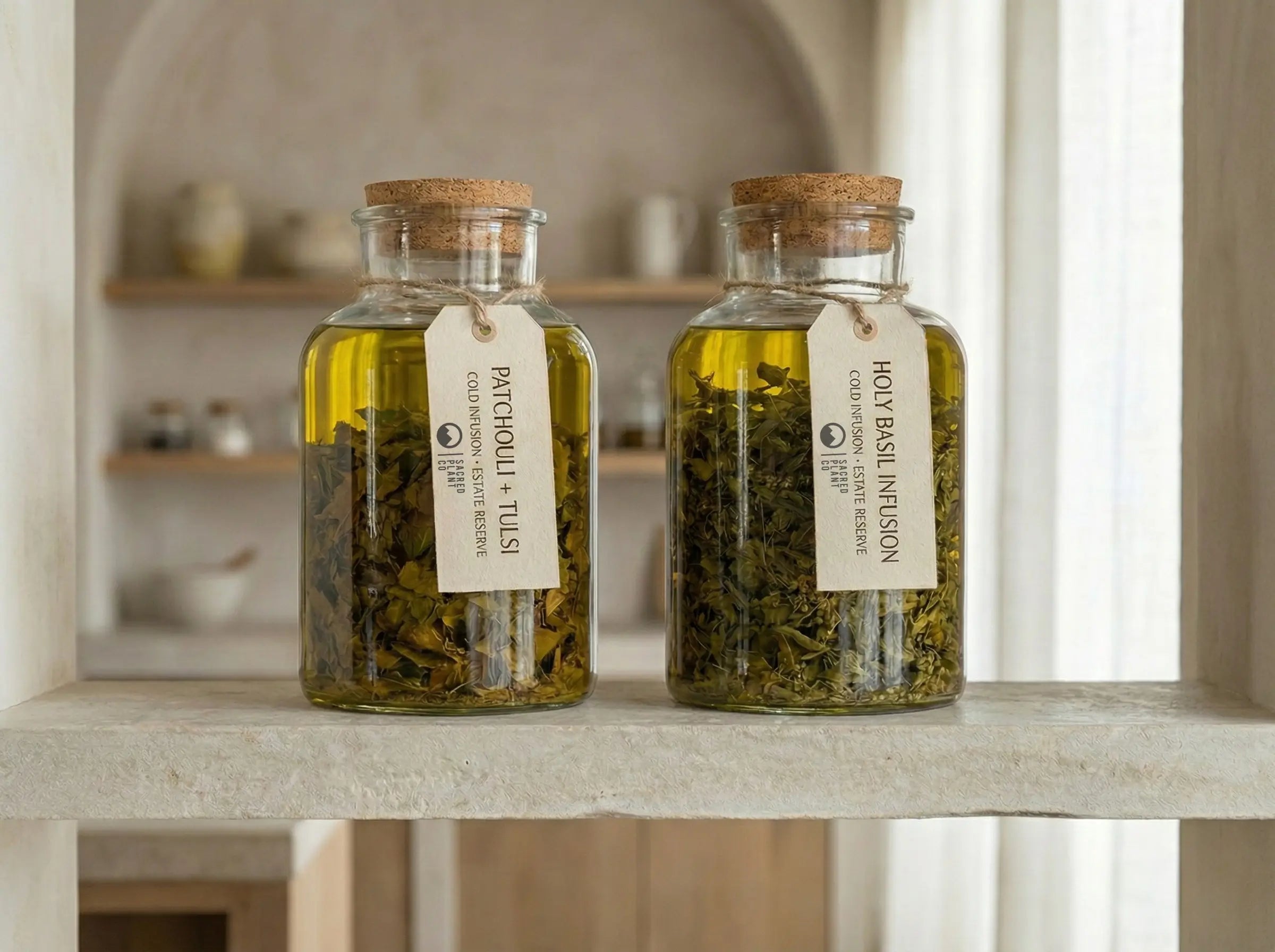Two glass apothecary jars on a limestone shelf containing a golden cold infusion of whole Patchouli and Tulsi leaves, labeled "Estate Reserve" and steeping in oil.