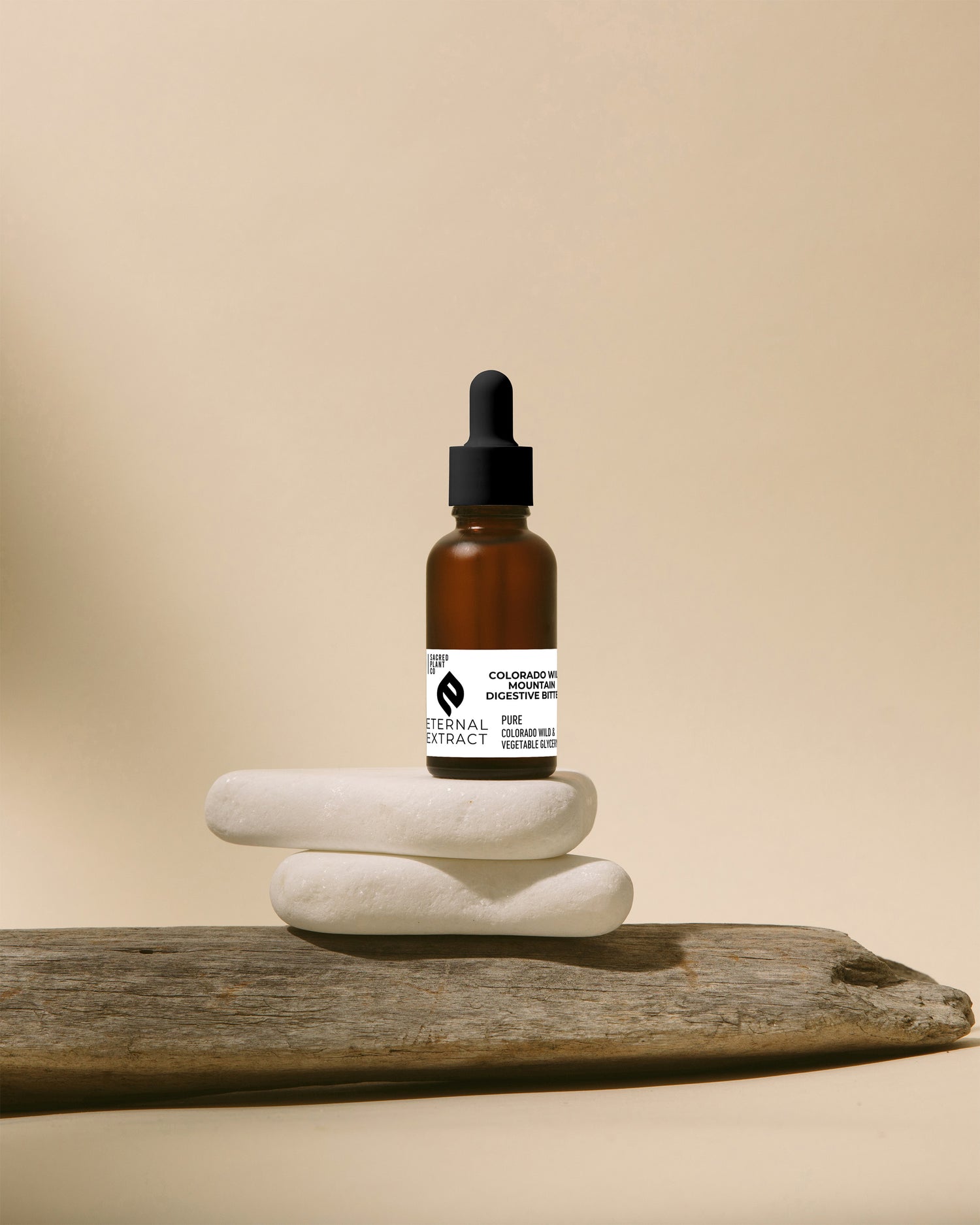 Sacred Plant Co Colorado Wild Mountain Digestive Bitters Eternal Extract in an amber glass bottle, balanced on stacked white stones with driftwood, against a calming beige background for a natural and earthy display.