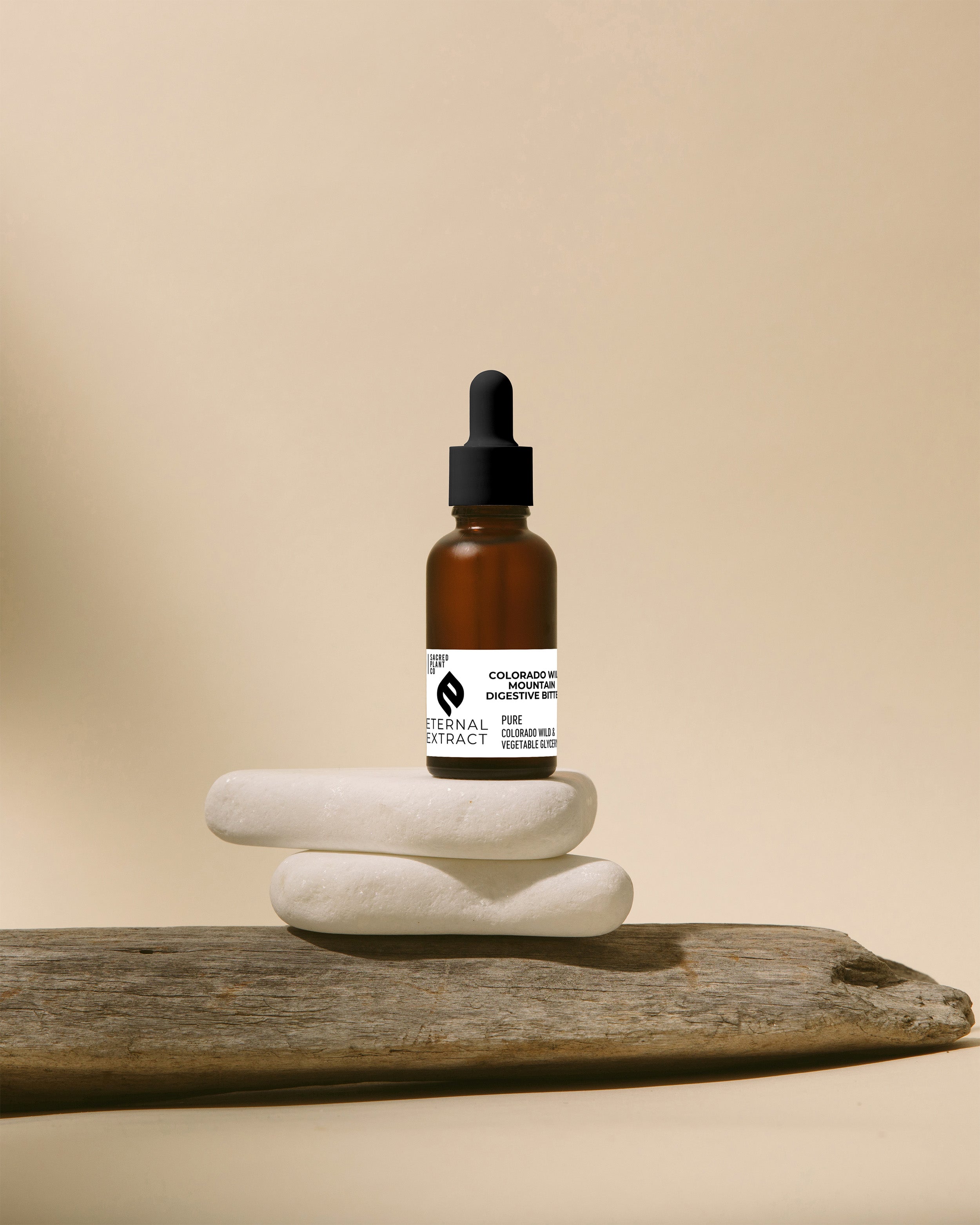 Sacred Plant Co Colorado Wild Mountain Digestive Bitters Eternal Extract in an amber glass bottle, balanced on stacked white stones with driftwood, against a calming beige background for a natural and earthy display.