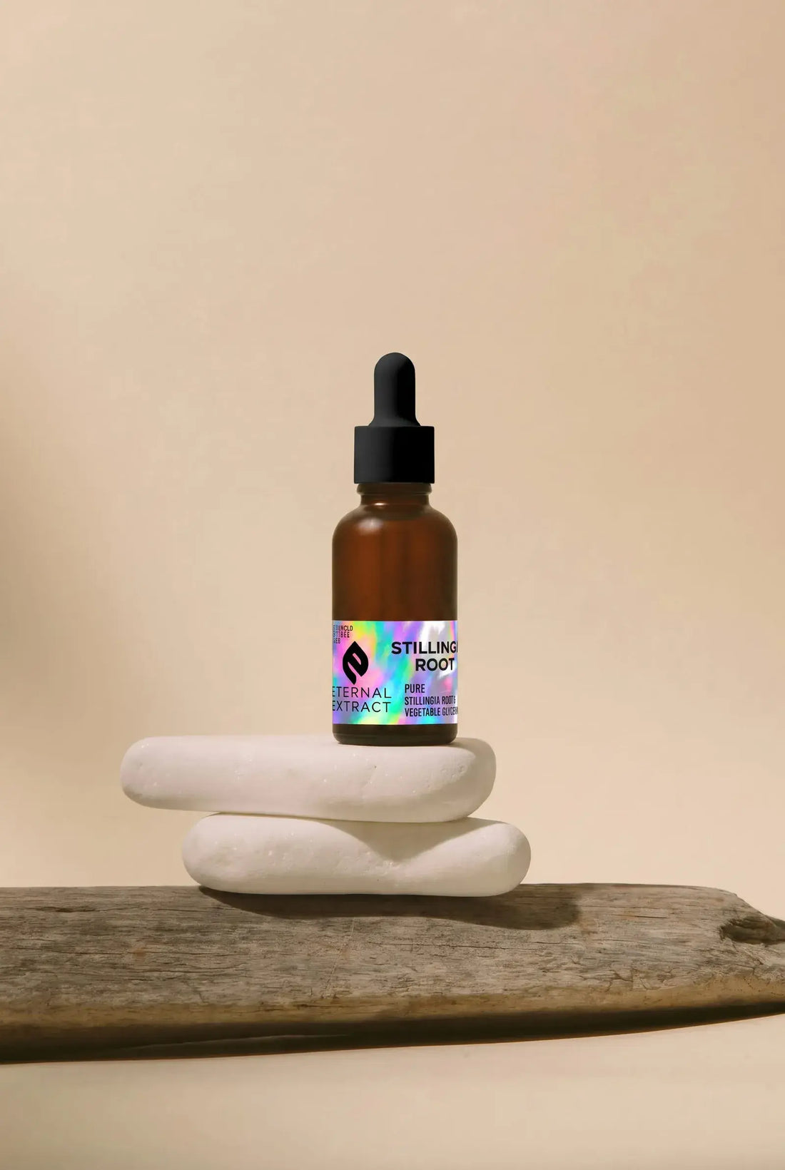 Sacred Plant Co Eternal Extract Stillingia Root tincture in an amber glass dropper bottle with a holographic foil label, balanced on two stacked white stones resting on weathered driftwood against a warm beige background.