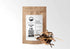 A brown paper bag of Stillingia Root from Sacred Plant Co. is shown against a white background. The bag has a white label with the company logo, product name, weight (4 OZ), a QR code, and tasting notes of straw, dandelion, and marshmallow root. Beside the bag is a small metal bowl overflowing with pieces of dried root.