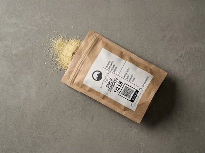 Sacred Plant Co. kraft paper pouch of Garlic Granules (1/2 LB) spilled onto a grey surface to show the coarse, golden-yellow texture. Label notes: Low Water Regenerative Colorado Farming.