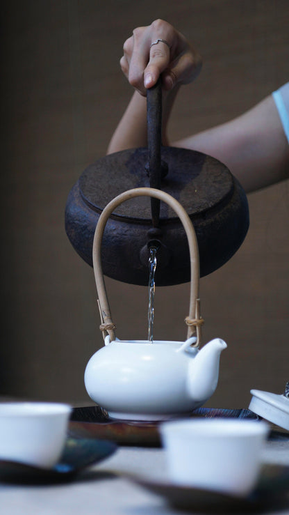 Pouring water from an antique iron kettle into a white teapot with a bamboo handle, traditional tea ceremony setup, close-up on hands.