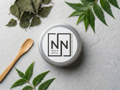 Neem &amp; Nettle salve tin labeled "NN" arranged on a grey surface with a wooden applicator spoon and dried herbal ingredients.