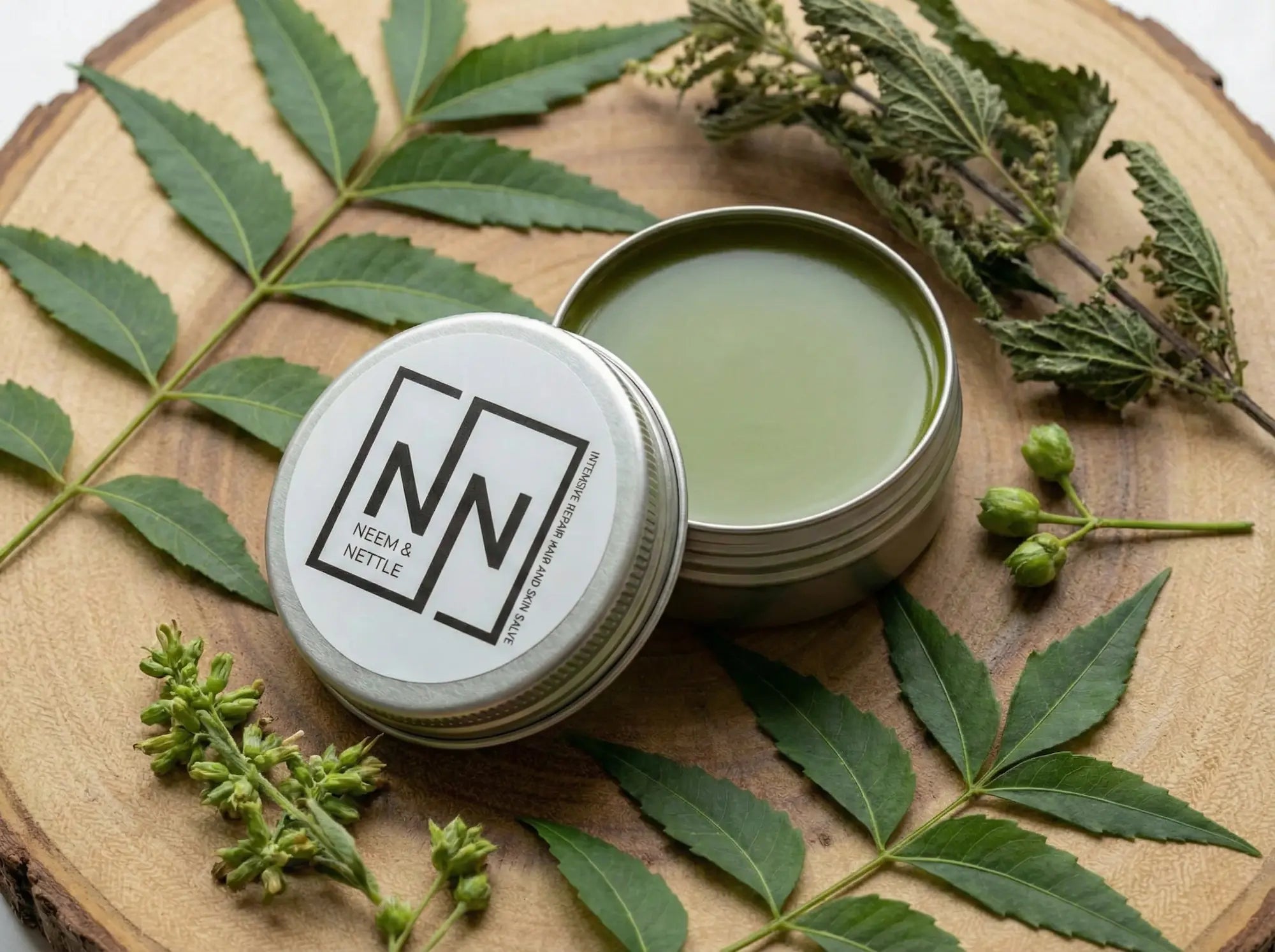 Open tin displaying the deep chlorophyll-green color of the Neem &amp; Nettle salve, resting on a wood slice amidst raw botanical ingredients.