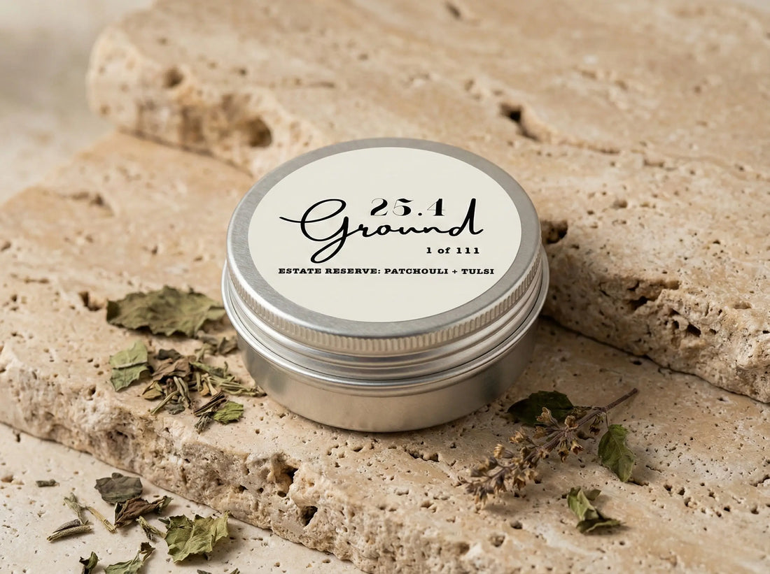 A closed silver tin of 25.4 Ground Estate Reserve Balm sitting on porous travertine stone, surrounded by dried patchouli and tulsi leaves.