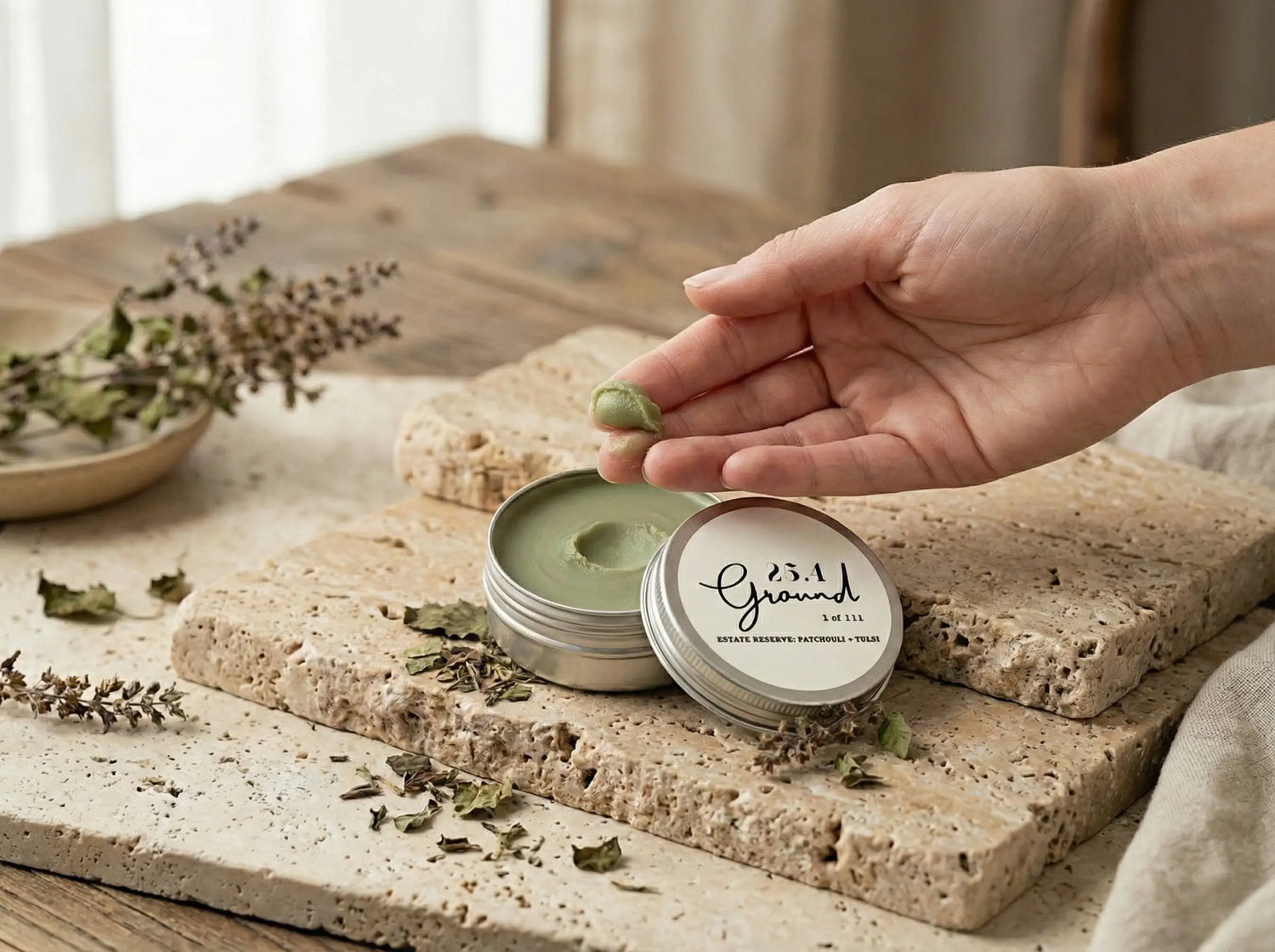 A hand scoops a pearl-sized amount of green herbal balm from a tin, demonstrating the smooth, melt-on-contact texture of the cold-infused oils.