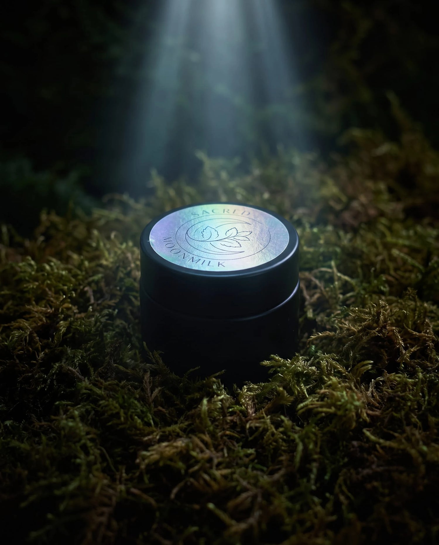 Sealed jar of Moonmilk Botanical Night Balm with holographic moon-and-botanical lid, standing on forest moss in a soft beam of light