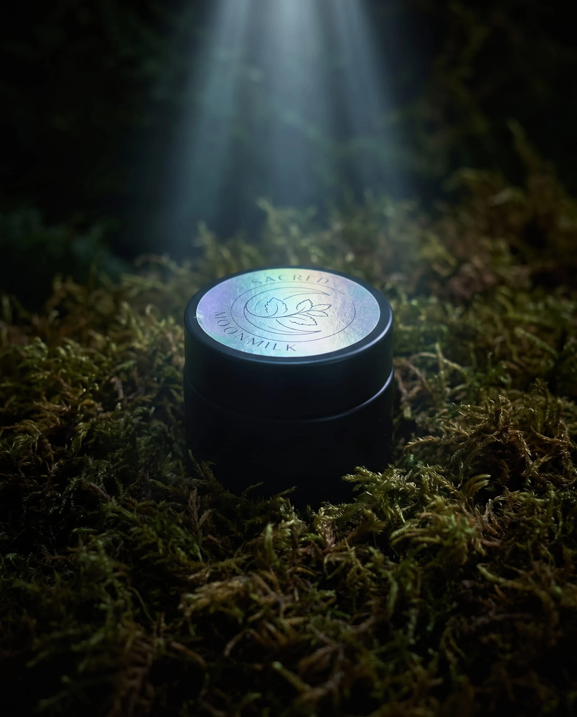 Sealed jar of Moonmilk Botanical Night Balm with holographic moon-and-botanical lid, standing on forest moss in a soft beam of light