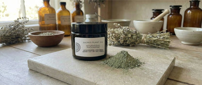 A dark amber glass jar of Sacred Plant Co Ayurvedic Face Mask sits next to a pile of raw green neem and clay powder, framed by dried lavender bundles in a sunlit herbal apothecary.