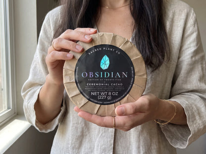 Woman in linen holding the 13 cm Obsidian Edition ceremonial cacao round in both hands beside a sunlit window