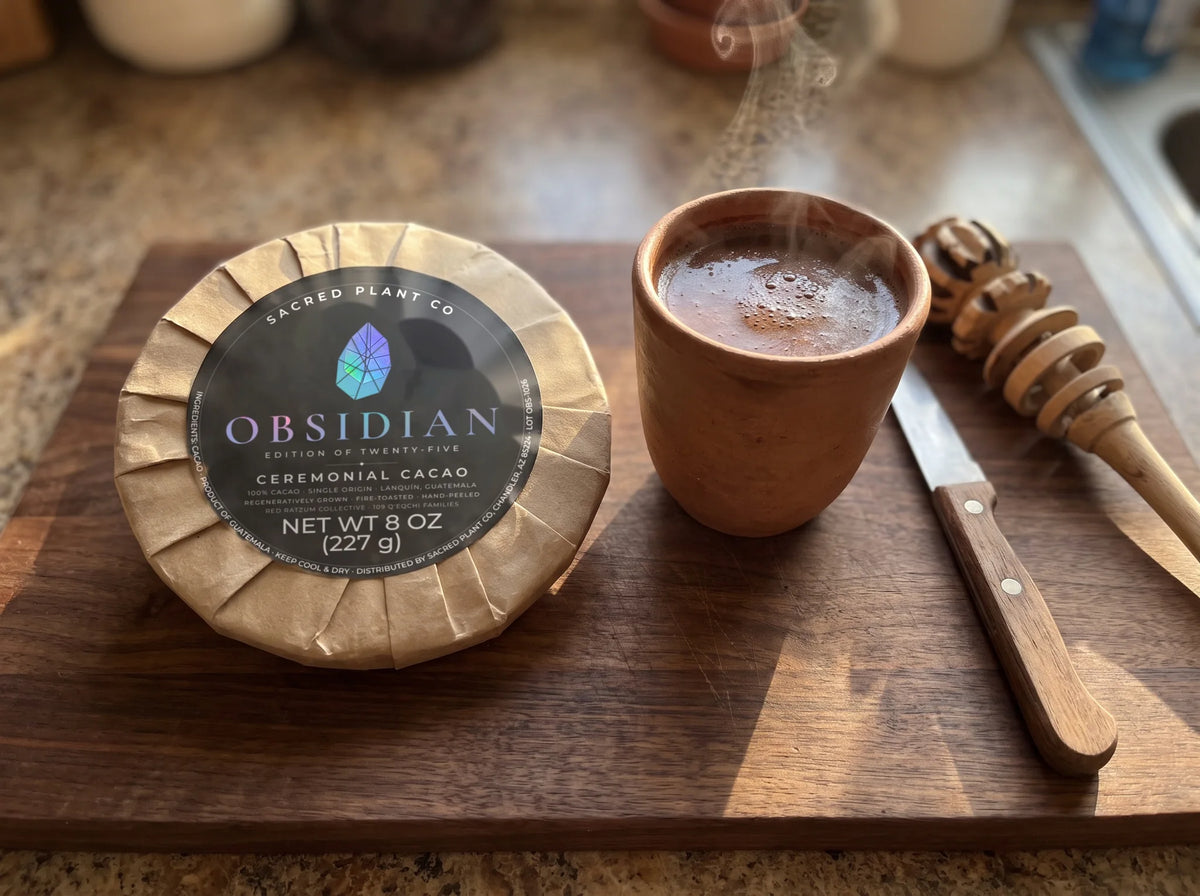 Steaming clay cup of ceremonial cacao on a walnut board beside the Obsidian round, a knife for shaving and a wooden molinillo