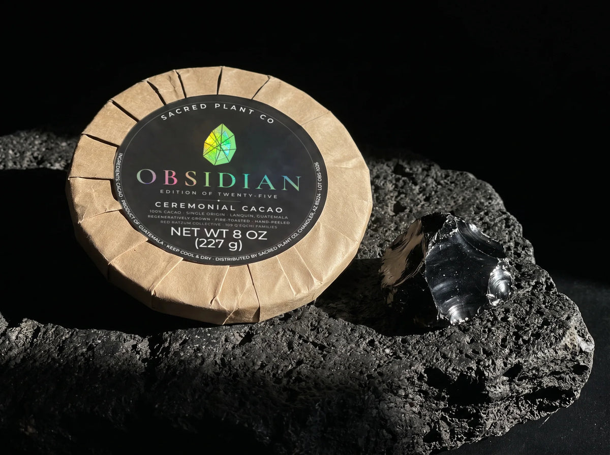 Obsidian Edition ceremonial cacao round on black volcanic rock beside a piece of raw obsidian, the stone the edition is named for