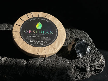 Obsidian Edition ceremonial cacao round on black volcanic rock beside a piece of raw obsidian, the stone the edition is named for