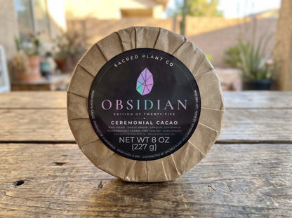Sacred Plant Co ceremonial cacao round standing on a weathered wood table outdoors, kraft pleats framing the black Obsidian label