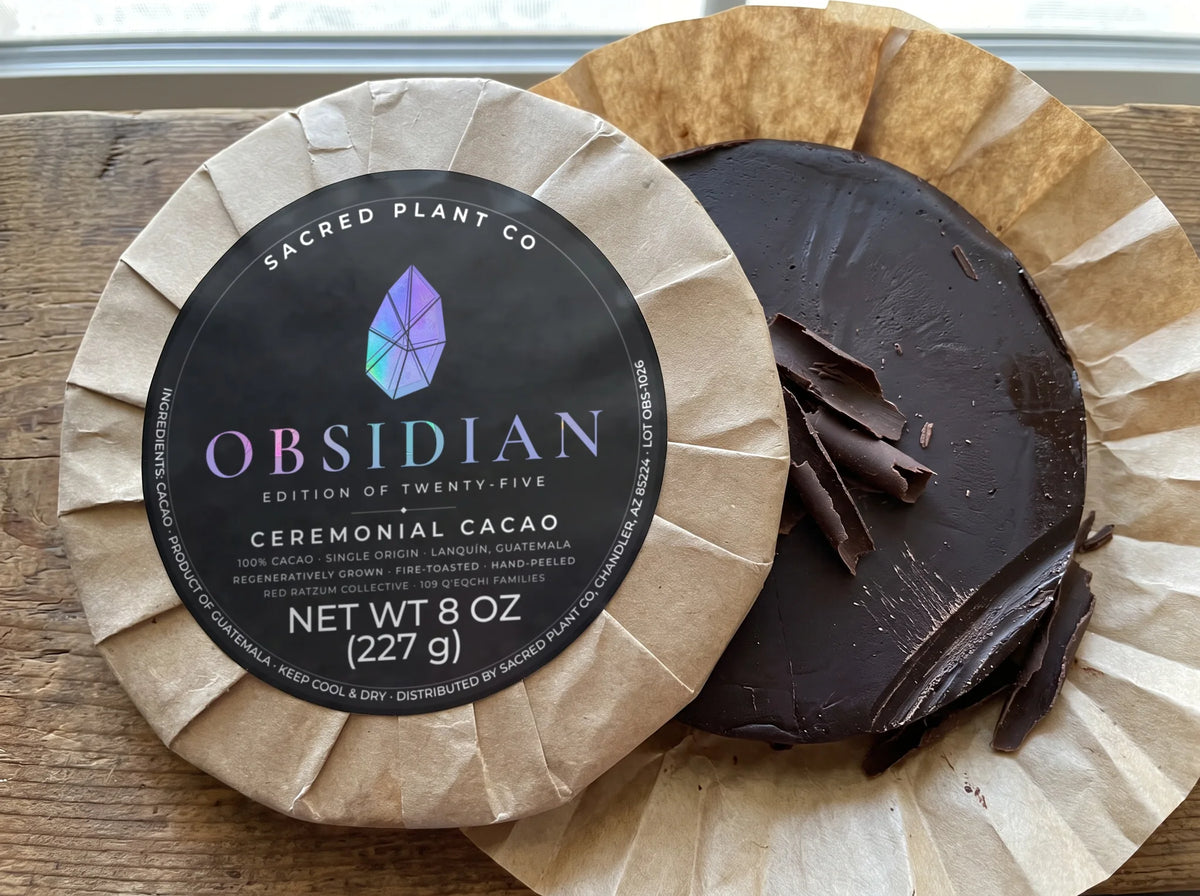 Unwrapped round of 100% cacao with dark shavings cut from the edge, behind a wrapped Obsidian Edition ceremonial cacao round
