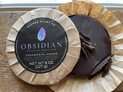 Unwrapped round of 100% cacao with dark shavings cut from the edge, behind a wrapped Obsidian Edition ceremonial cacao round