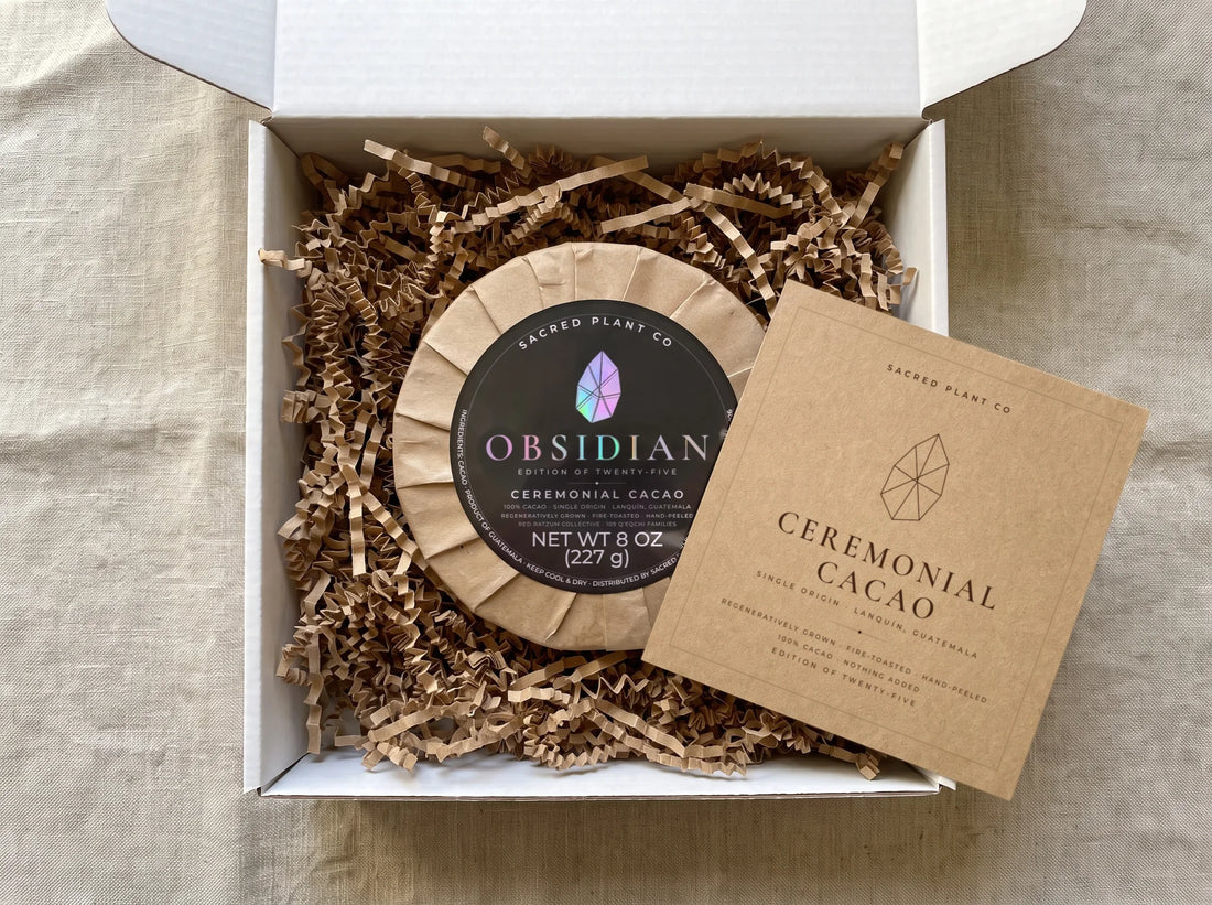 Ceremonial cacao gift box opened: the Obsidian round nested in kraft crinkle fill in a white mailer beside its kraft preparation card