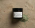 A flat-lay photo of the Sacred Plant Co Ayurvedic Face Mask jar and a dusting of green clay powder on a rustic concrete background.