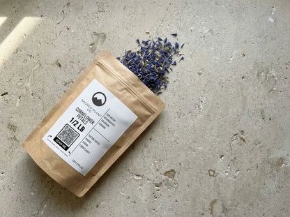 Sacred Plant Co Cornflower Petals 1/2 LB kraft paper bag lying flat on a textured stone surface, with dried blue petals spilling naturally from the top