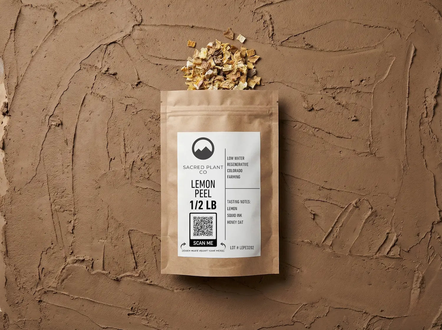 Sacred Plant Co 1/2 lb bag of dried lemon peel lying flat on a textured clay surface. Small pieces of dried golden lemon peel spill naturally from the top of the kraft paper packaging, which features a label reading &