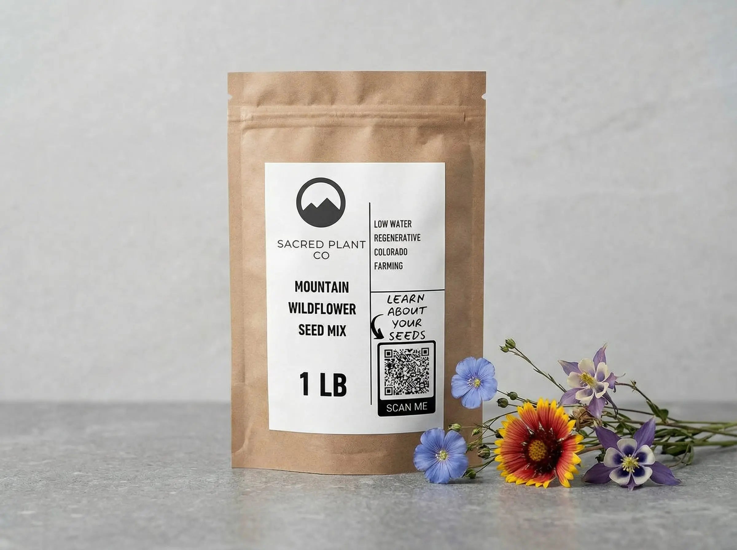 Sacred Plant Co Mountain Wildflower Seed Mix pouch displayed on stone with fresh blooms of blue flax, blanketflower, and columbine in a clean studio setting.