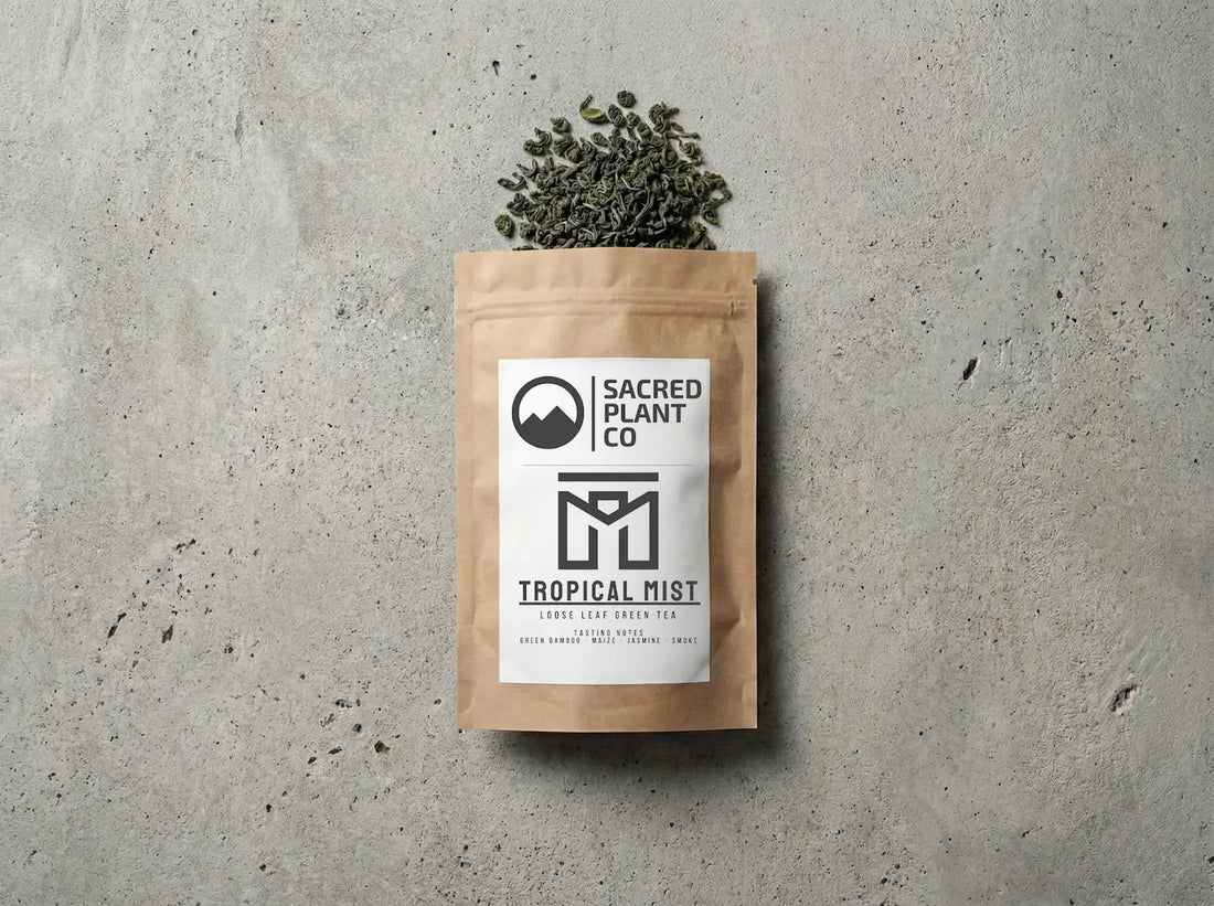 Sacred Plant Co. Tropical Mist loose leaf green tea in a brown kraft bag, layflat editorial photography on a textured stone surface. Premium Chun Mei style green tea leaves with tasting notes of green bamboo, maize, jasmine, and smoke spilling naturally from the packaging.