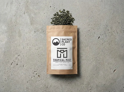 Sacred Plant Co. Tropical Mist loose leaf green tea in a brown kraft bag, layflat editorial photography on a textured stone surface. Premium Chun Mei style green tea leaves with tasting notes of green bamboo, maize, jasmine, and smoke spilling naturally from the packaging.