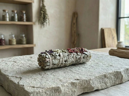 White sage smudge stick displayed on a stone slab in an herbal apothecary setting with jars in the background.