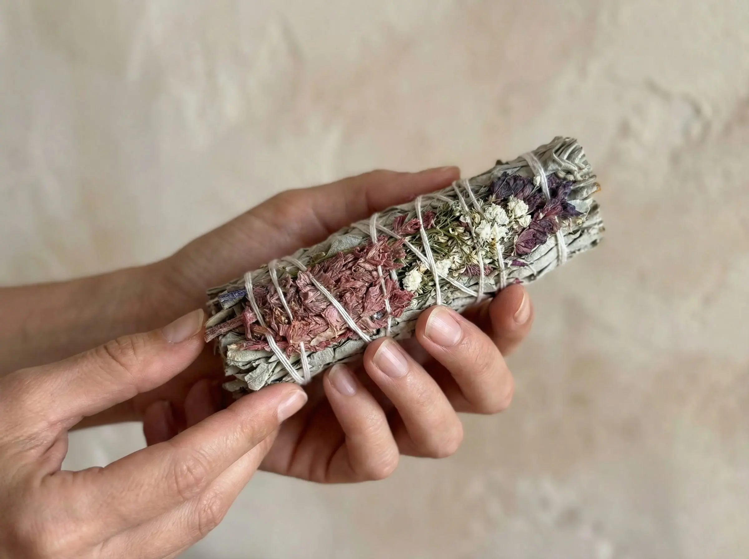Close-up of a hand-tied white sage smudge stick held in hands, showing leaf texture and dried florals.