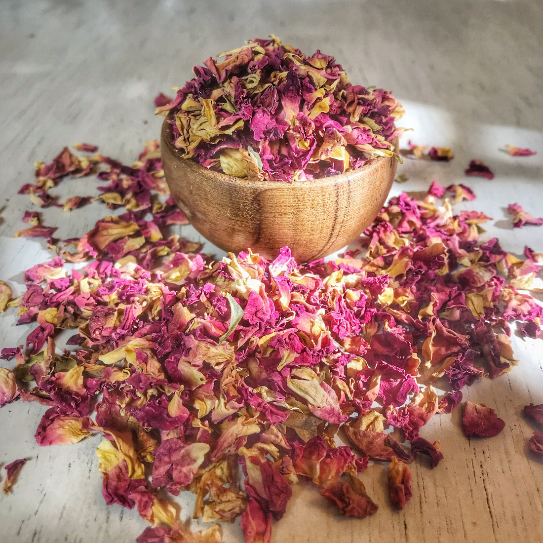 Organic dried rose petals in a natural wooden bowl, spread on a wooden surface, suitable for herbal teas and crafts.