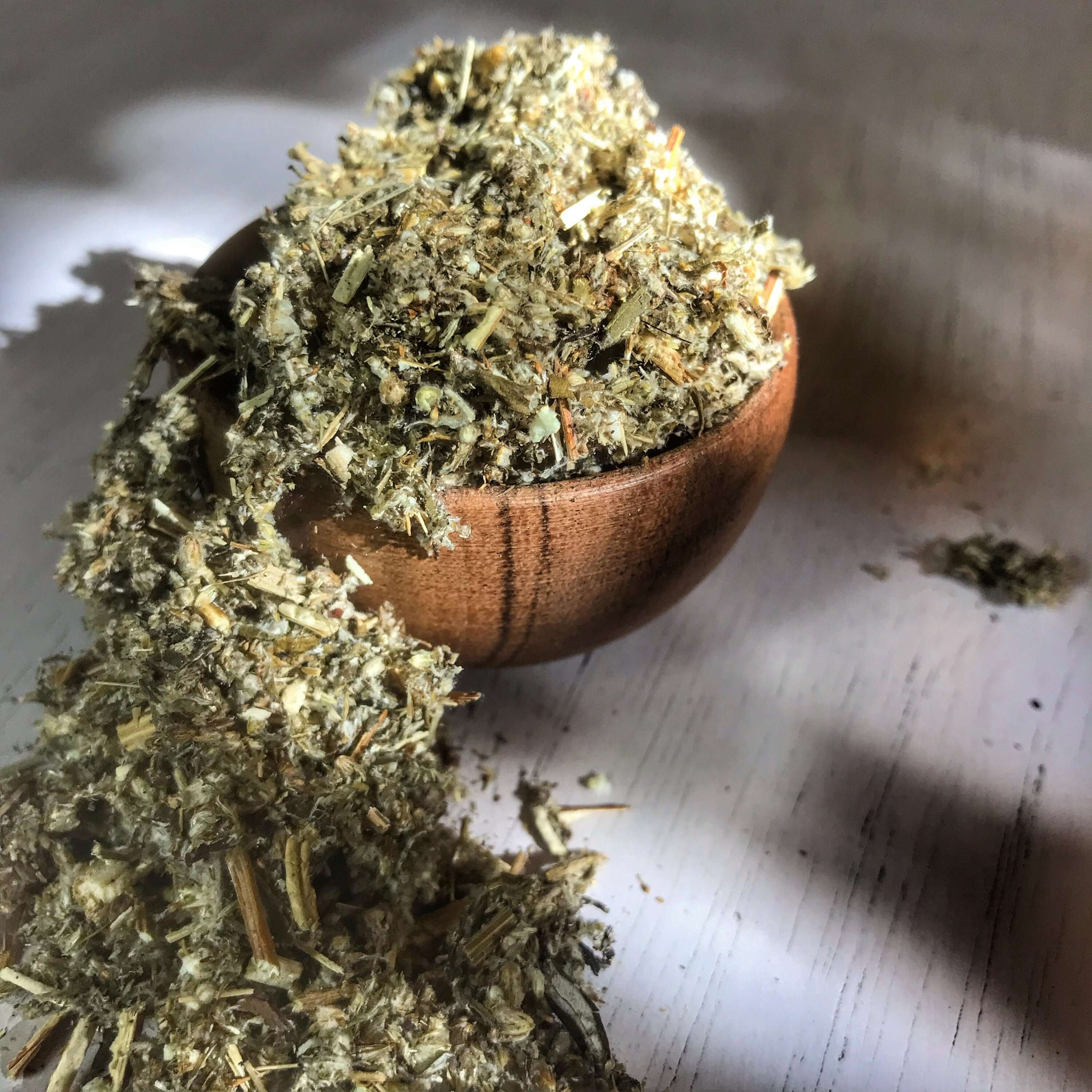 Mugwort Herb tumbling from a wooden bowl, captured in soft light, representing the earthy texture and superior quality of Sacred Plant Co&