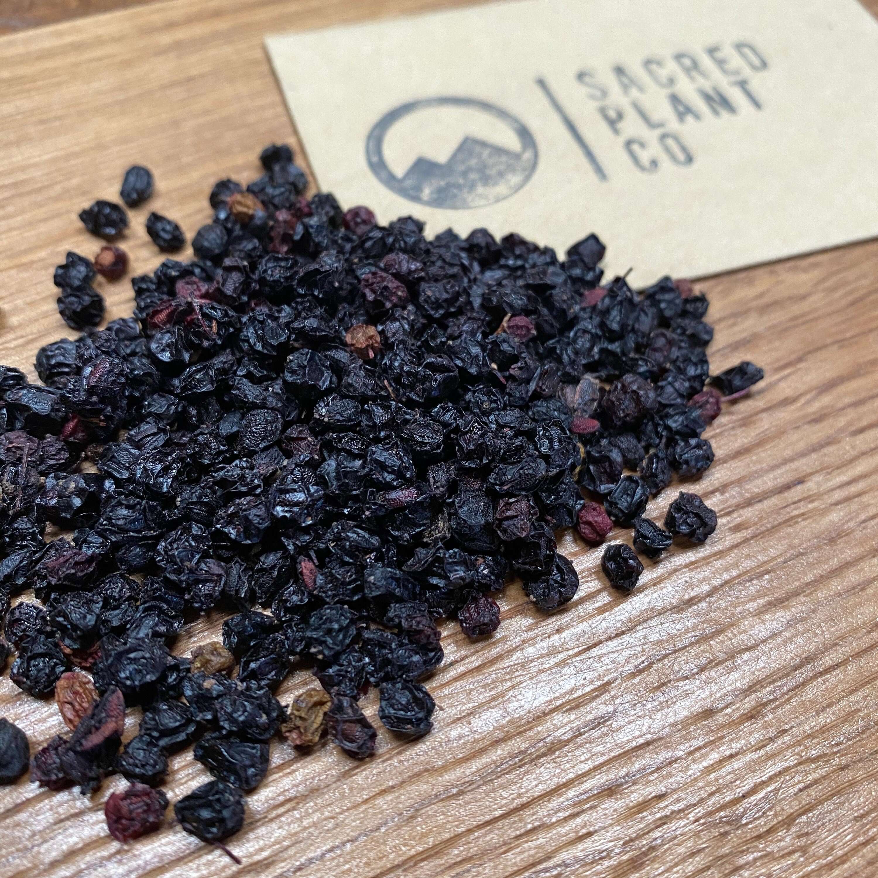 Close-up of dark, wrinkled Whole Elderberries, texture contrasted with wooden backdrop, showcasing Sacred Plant Co’s premium herbs.