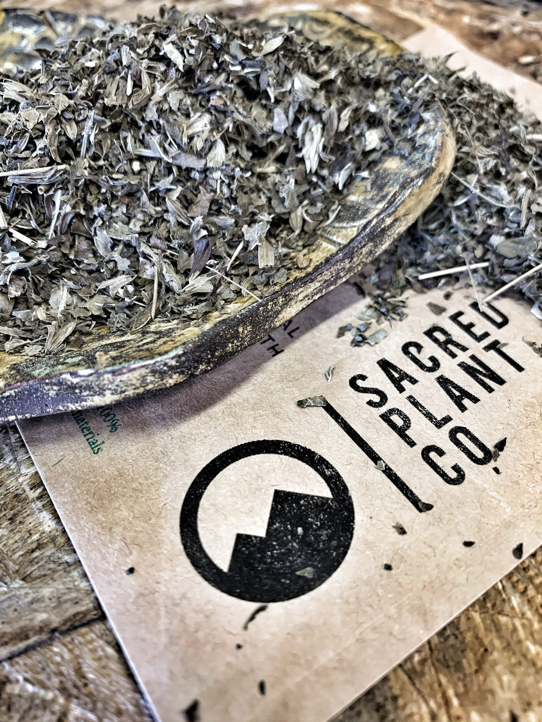 Close-up of dried lemon balm leaves on a rustic wooden bowl over a card with the Sacred Plant Co logo, accentuating the finely chopped texture and natural quality of the product.